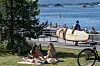 Folk soler seg på gresset ved vannet på Sørenga i Oslo, med strand, brygge og båter i bakgrunnen.