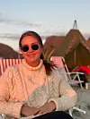 Kvinne i hvit strikkegenser og solbriller sitter i en strandstol med telt i bakgrunnen.