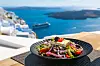 Bilde av en gresk salat med oliven, rødløk, tomater og fetaost, med Santorini i Hellas i bakgrunnen.