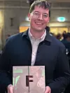 En mann med briller smiler og holder en pris med bokstaven F innendørs.