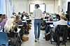Kvinnelig lærer underviser elever i et klasserom med digital tavle og bordplasser.