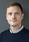 Portrett av en mann med lys skjorte og mørk genser foran grå bakgrunn.