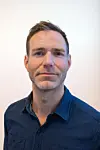Joakim Øverbø i blå skjorte foran en lys vegg.