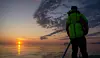 En person står i silhuett med grønn jakke ved et rolig hav i solnedgang.