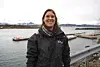 Ein person i svart jakke står ved ei kai med vatn og fjell i bakgrunnen.