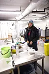 En person i laboratorium står ved et bord med prøver, bøtter og utstyr under store ventilasjonsrør.