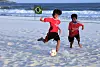 To gutter spiller fotball på en strand med havet som bakgrunn. Et brasiliansk flagg er satt opp i utkanten av der guttene spiller.