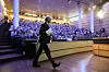 En mann i mørk dress går foran et stort publikum i et auditorium med trepanel og trappetrinn.