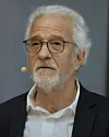 Eldre mann med briller og hvitt skjegg foran grå bakgrunn, med mikrofon ved kinnet.