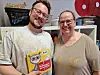 To personer smiler og holder en fargerik barnbok innendørs foran en hylle.