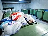 Overfylte grønne søppelkontainere og store avfallsposer i et parkerings- eller avfallsrom.