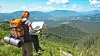 Person med ryggsekk sitter på en stubbe og holder et kart med fjell og skog i bakgrunnen.