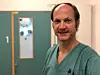 Portrettbilde av Knut Jørgen Labori i grønn sykehusskjorte. Han står i en sykehuskorridor. I bakgrunnen ses en grønn dør med et vindu, og innenfor der ser det ut som det er en operasjonssal der en operasjon er i gang.