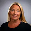 Portrett av en kvinne med langt blondt hår og mørk topp foran en grå bakgrunn.