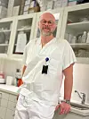 En mann i hvit uniform står i et lyst klinikkrom med hyller, flasker og vask i bakgrunnen.