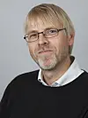 Portrettbilde av insektsforsker Hallvard Elven. Han har kort lyst hår og briller, litt skjegg som begynner å gråne, en svart genser og en hvit skjortekrage som stikker opp rundt halsen.