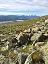 Steinete fjellside med gress, innsjø og fjell i bakgrunnen.