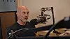 En skallet mann sitter i et radiostudio foran en mikrofon og mikrofonarm.