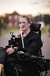 Kvinne i rullestol med pustemaske og svart utstyr foran seg, fotografert utendørs i varmt kveldlys.