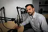 En mann sitter ved et bord med mikrofoner og hodetelefoner i et lite podkaststudio.