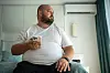 Overvektig mann med skjegg sitter på en seng og ser på mobiltelefonen i et lyst soverom.