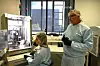 To personer i verneklær arbeider ved en arbeidsbenk med laboratorieutstyr og en skjerm i et forskningsrom.