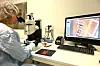 Person i vernedrakt arbeider ved mikroskop på et laboratorium med dataskjerm og utstyr.