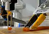 To industriroboter arbeider ved et bord med oransje plastdeler i et laboratorie- eller fabrikklokale.
