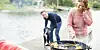 To personer står ved en grill på en brygge ved vannet, med mat og utstyr på bordet.
