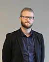 En mann med briller og skjegg står foran en grå vegg iført mørk blazer og skjorte.