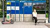 Kvinne sitter på en benk ved en bussholdeplass mens en el-sparkesykkel står ved siden av.