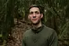 En mann står foran en tett skog med grønne trær og dempet lys.