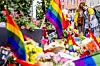 Blomster, lys og regnbueflagg ligger samlet ved et minnested utendørs.
