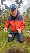Bilde av Arne Håkon Thomassen på feltarbeid. Han sitter på knærne og skal til å putte noe jord eller en gjenstand i en liten plastpose. Han har briller og skjegg, og en caps på hodet. Han har på mørke regnbukser og en rød og blå allværsjakke.