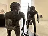 To mørke bronsefigurer i løpestilling står på sokler i et lyst museumsgalleri.