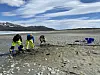 Tre arkeologer står og graver i sjøbunnen til innsjøen Tesse. I bakgrunnen ses fjell, blå himmel og skyer.