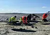 Fire arkeologer graver i sjøbunnen i Tesse. Vi ser et stort hull helt foran. I bakgrunnen er himmelen blå.