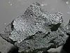 Et mørkt, uregelmessig meteorittfragment med sølvaktig, ru overflate på grå bakgrunn.