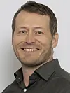 Nærbilde av en smilende mann i mørk skjorte foran lys bakgrunn.