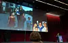 Scene med stor skjermprojeksjon, røde gardiner og talerstol under en presentasjon.