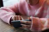 En ung jente med en mobiltelefon. Vi ser bare halve ansiktet hennes, hun sitter ved et bord og scroller på mobilen sin, iført en rosa genser.