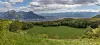 Bredt landskap med grønn dal, trær, fjell og skyet himmel. På gressletten synes en ring av grønnere gress.
