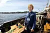 Person standing on a wooden boat deck with ropes and equipment on calm water near shore.