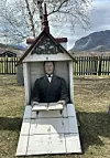 Lite kapellformet gravminne med skulptur og åpen bok på en gressplen foran fjell og gjerde.