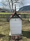 Liten hvit gravstøtte med rødt tak står i gresset foran et gjerde og fjell i bakgrunnen.