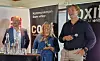 Tre personer står på en konferansescene med mikrofoner og et banner med tekst og logo i bakgrunnen.