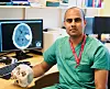 Portrettbilde av Sasha Gulati. Han har på seg grønne sykehusklær og holder en halv hodeskalle i hånda. I bakgrunnen skimtes en datamaskin med bilde av et tverrsnitt av en hjerne.