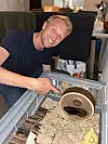 Smilende mann ved et trådbur med strø, matskål og en hamster i løpehjul.
