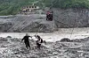 To personer krysser en steinete, flomrammet elv mens en ziplinekurv henger over vannet i Nepal.