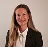 Portrett av en smilende kvinne med langt hår, hvit skjorte og mørk blazer mot en lys bakgrunn.